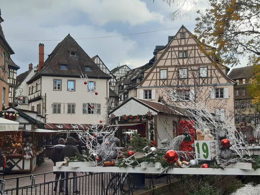 Festive atmosphere at the Christmas market in Colmar, France, with half-timbered houses adorned with seasonal decorations, shoppers browsing holiday stalls, and a large advent calendar adding to the quaint charm of the town