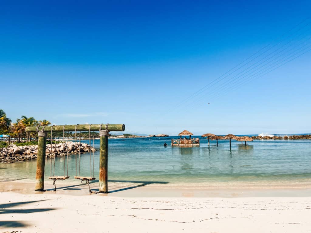A tranquil beach scene with clear blue skies and calm waters, featuring a wooden swing set on the sandy shore in the foreground and a charming floating bar surrounded by rocks in the distance, under the serene embrace of palm trees.
