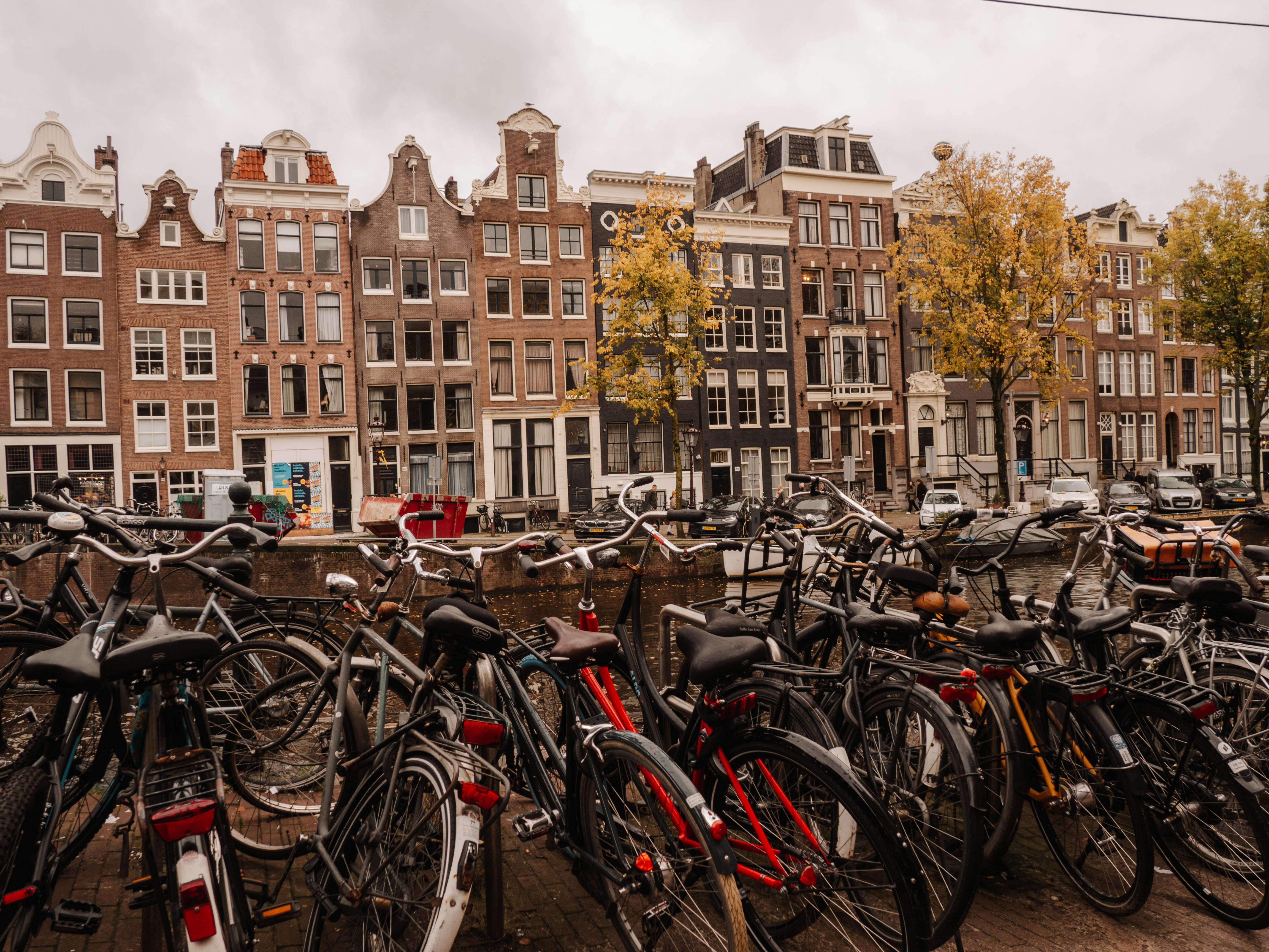 A typical canal-side view in Amsterdam, Netherlands, with a foreground of numerous parked bicycles and a backdrop of historic gabled houses, reflecting the city's cycling culture and distinctive architectural style on an overcast day.