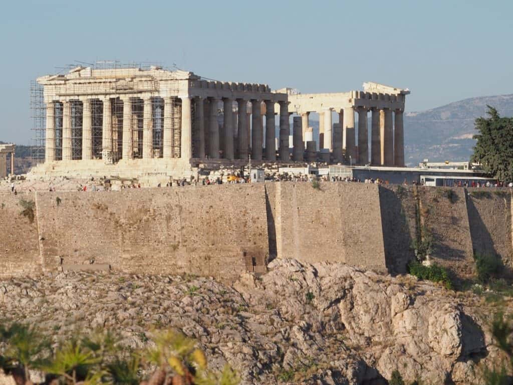 The Parthenon, the iconic ancient temple on the Acropolis of Athens, Greece, stands against a backdrop of mountains, its Doric columns and partially restored facade bathed in the warm glow of the setting sun.