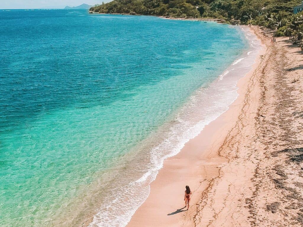A solitary figure walking along the pristine shoreline of Camp Bay Beach, Roatan, with the clear turquoise Caribbean Sea on one side and a line of lush vegetation on the other, under a bright tropical sun.
