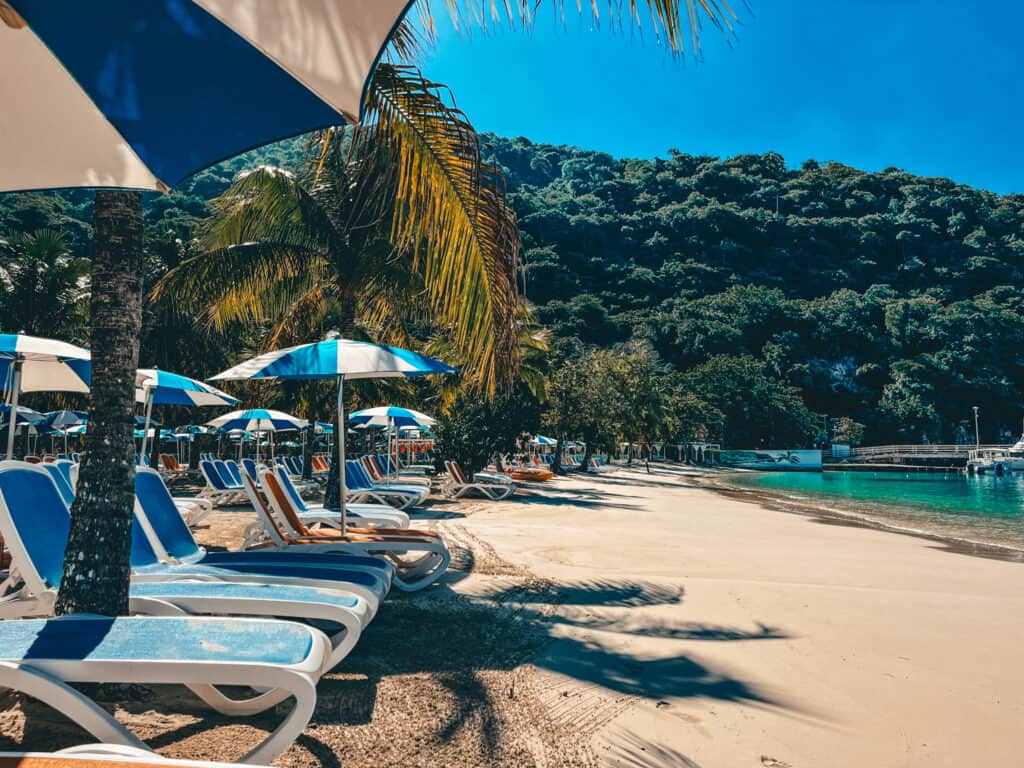 A picturesque view from the shade under a beach umbrella, showing a row of white and blue sun loungers on the sandy shore, with tropical palm trees and a dense hillside of verdant foliage in the background, beside a tranquil turquoise sea.
