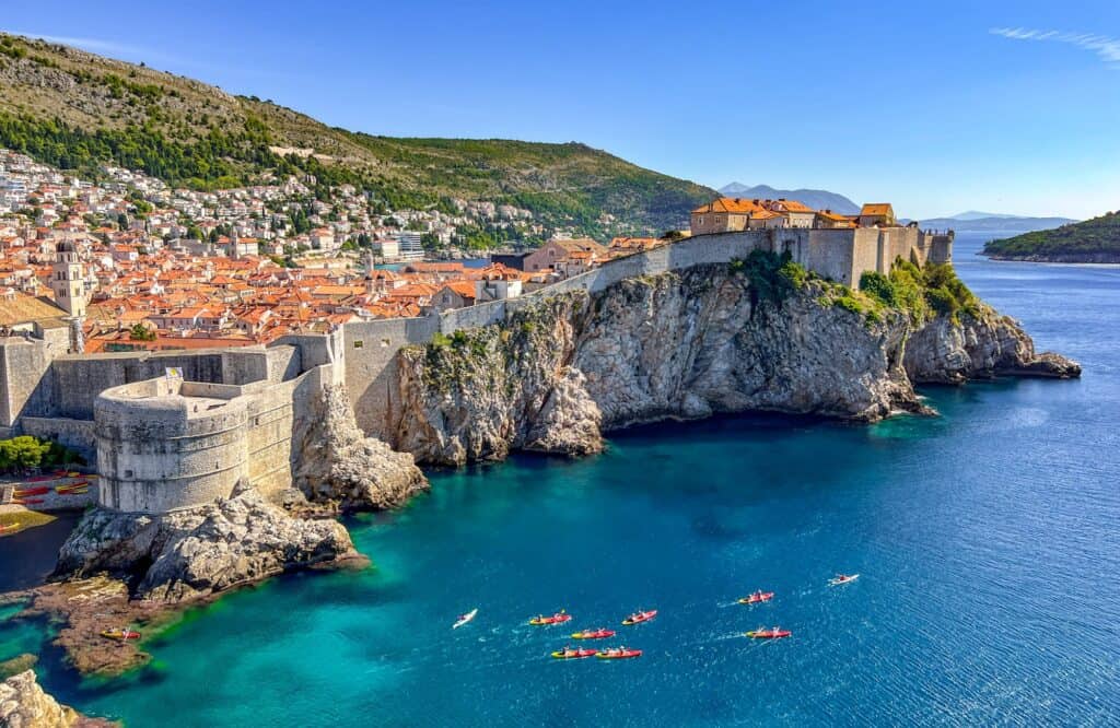 Aerial view of Dubrovnik's old city walls and fortifications extending into the Adriatic Sea, with kayakers paddling in the clear blue water below and the terracotta-roofed cityscape backed by a verdant hillside.