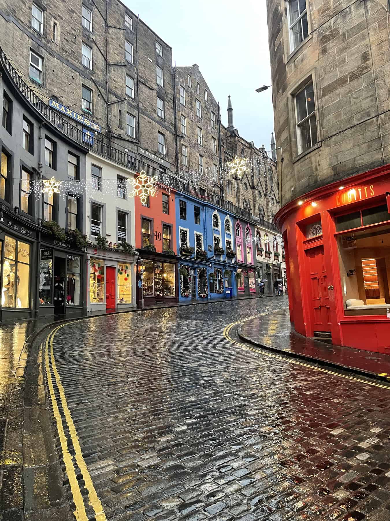 A vibrant street in Edinburgh, Scotland, with colorful shopfronts and holiday decorations reflecting off the wet cobblestones after a rain, creating a festive and cozy atmosphere in the historic city