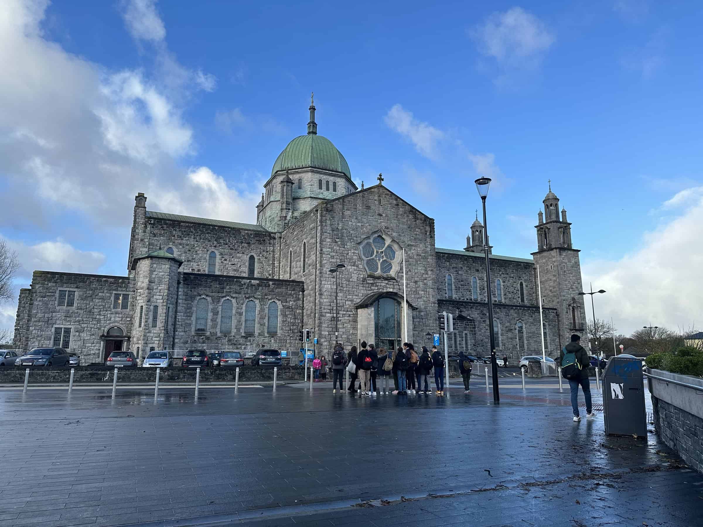 The Galway Cathedral with its impressive stone architecture and green dome stands under a partly cloudy sky, as pedestrians walk by the wet pavement reflecting the bright, scattered clouds after a rain shower in Galway, Ireland.