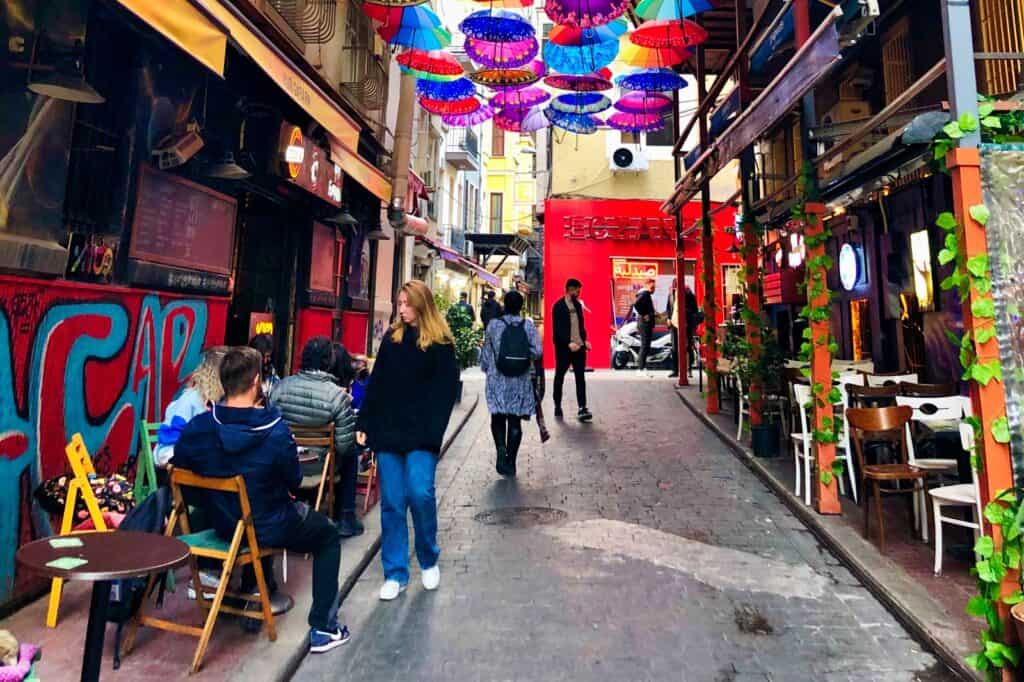 A lively alley in Istanbul, Turkey, lined with colorful umbrella art installations above and vibrant cafes on either side, while pedestrians enjoy the bohemian and artistic vibe of the city