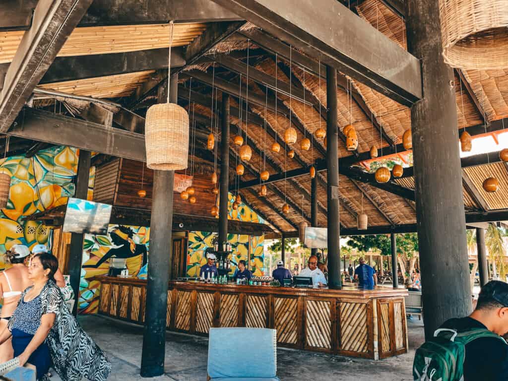 A lively beach bar with a rustic charm, featuring a thatched roof, bamboo accents, and unique hanging lanterns, with patrons enjoying the ambiance and colorful mural art, creating a cozy tropical atmosphere.
