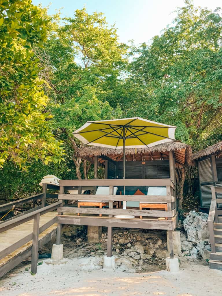 A secluded beach cabana with a yellow umbrella, offering a private slice of paradise with comfortable seating, nestled among lush greenery and rugged rocks, providing a perfect spot for relaxation by the sea.