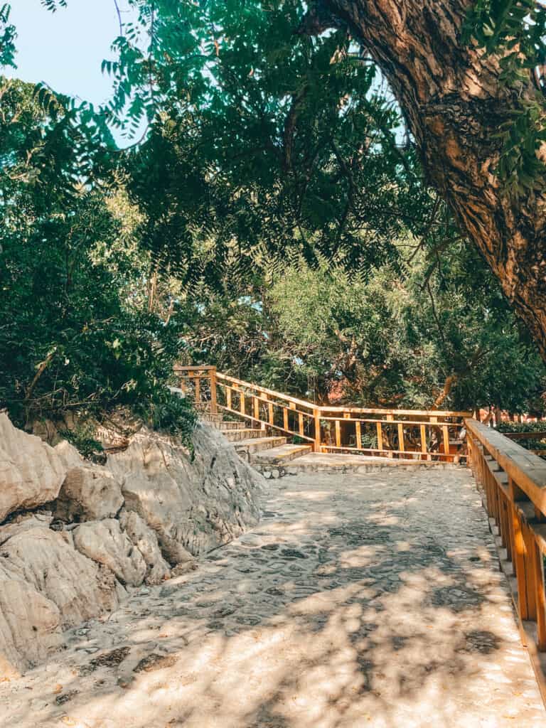 A serene nature path shaded by leafy green trees, leading to a wooden footbridge with a railing, over rugged terrain and white rocks, inviting a walk through a tranquil, sun-dappled environment.