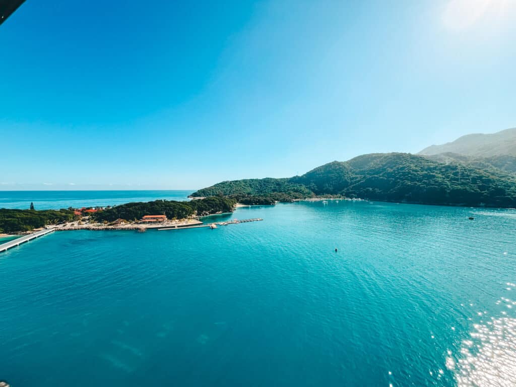 GPT An aerial view of a serene port in Labadee, showcasing the clear blue waters of the Caribbean Sea, with a dock extending into the water, surrounded by lush green hills and a tranquil beachfront, under a bright blue sky.