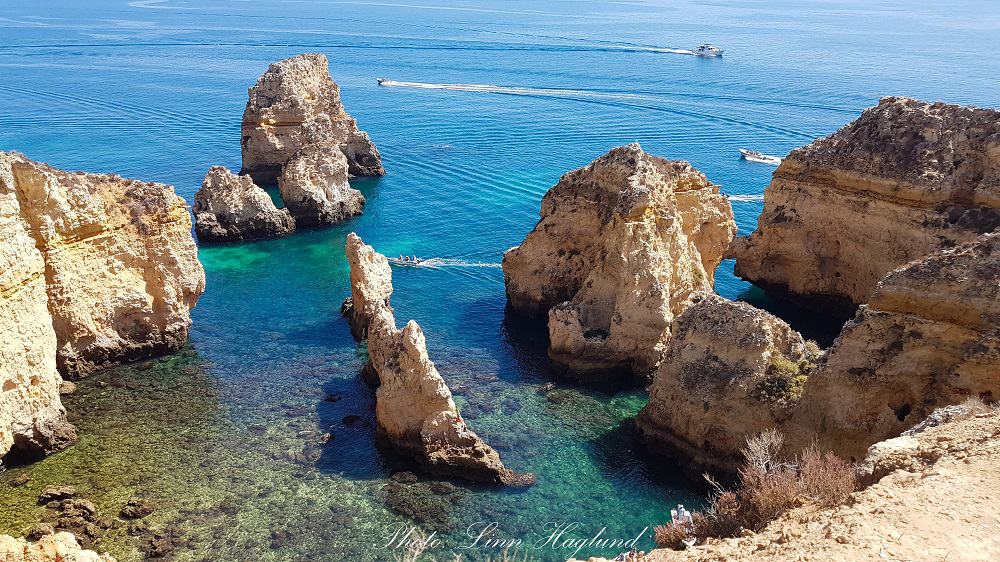 Crystal-clear turquoise waters surround the rugged limestone rock formations off the coast of Lagos, Portugal, with leisure boats cruising by, capturing the essence of the Algarve's stunning natural beauty