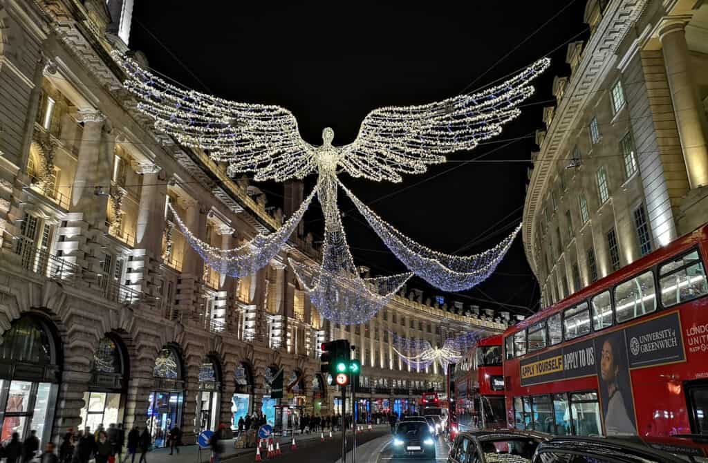 Nighttime view of Regent Street in London adorned with festive holiday lights, featuring an illuminated angel-like figure with widespread wings, with busy street traffic including the iconic red double-decker buses below.