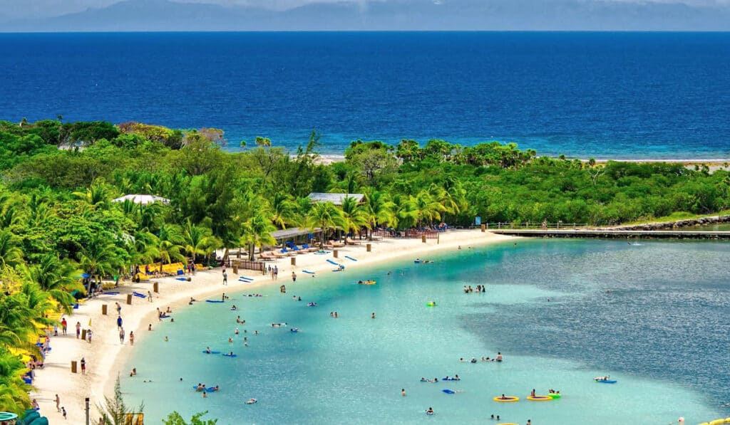 Vibrant view of Mahogany Beach in Roatan, showcasing people enjoying the calm turquoise waters, sandy shorelines, and recreational activities, set against a backdrop of lush tropical foliage and the expansive Caribbean Sea.