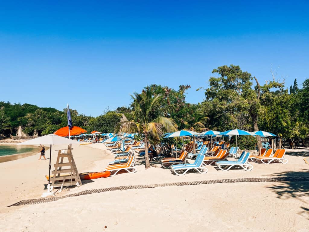 A vibrant beachfront lined with brightly colored sun loungers and umbrellas on golden sands, with lush greenery in the background and a lifeguard stand to the left, under a cloudless blue sky.