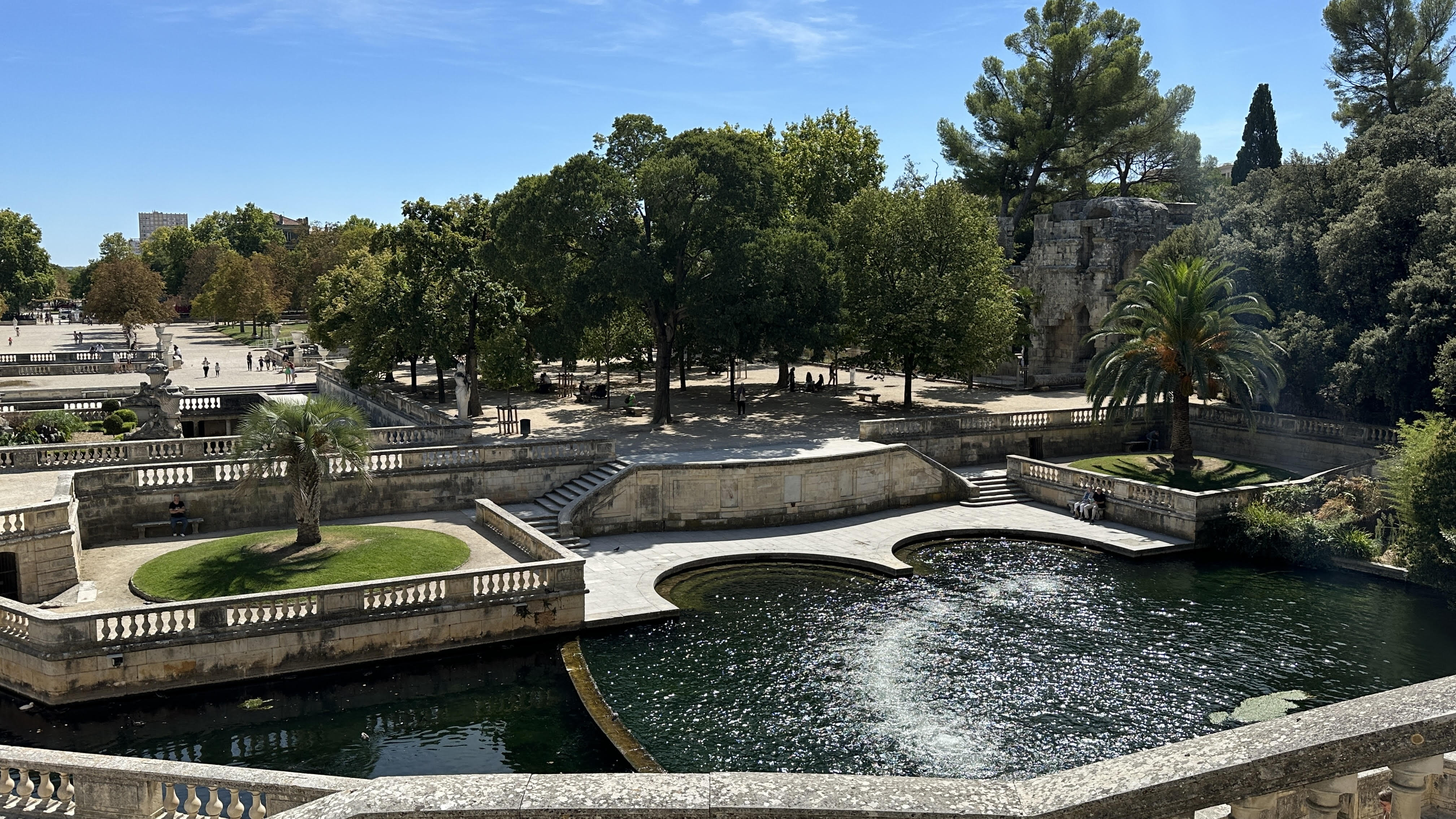 Scenic view of the Jardins de la Fontaine in Nîmes, France, featuring terraced gardens with classical stone balustrades, lush greenery, and a glistening pond, with visitors enjoying the sunny ambiance.