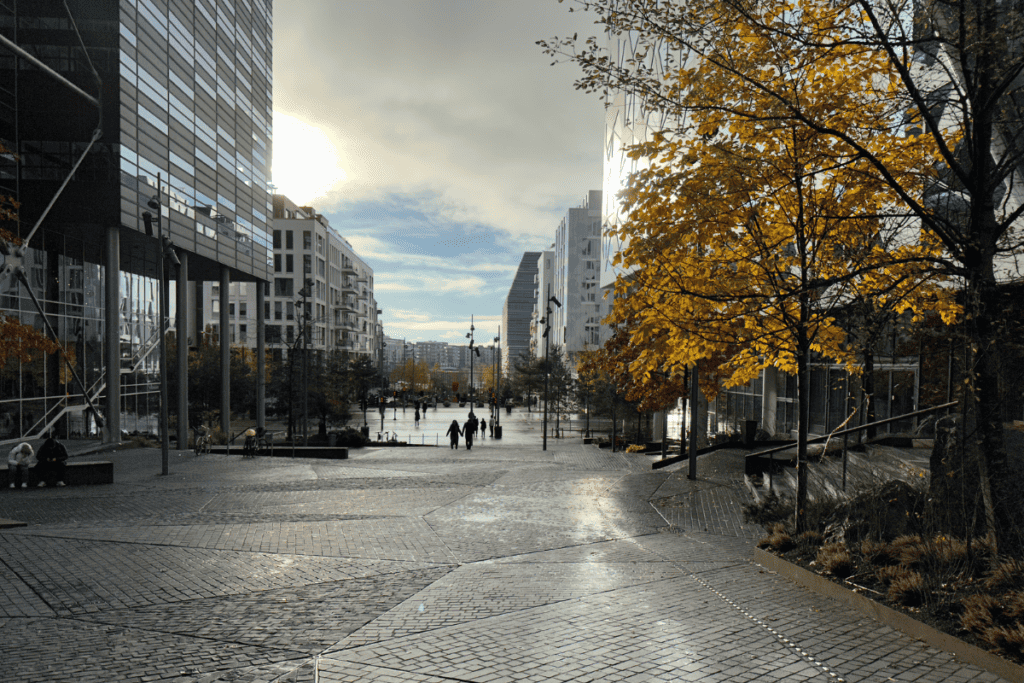 A wet urban plaza in Oslo, Norway, glistens under the autumn sun with golden leaves adorning trees, as pedestrians walk between modern buildings reflecting the bright sky.