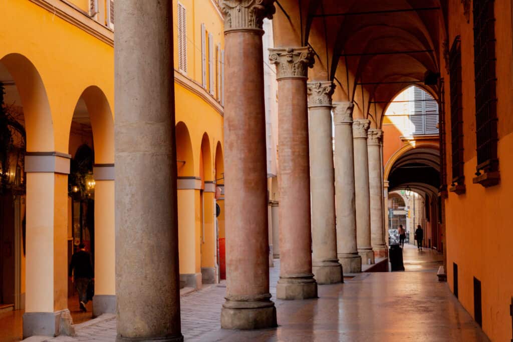 Warmly lit porticoes in Bologna, Italy, with a succession of elegant columns and arches casting soft shadows on the pavement, as locals stroll through this historic architectural feature typical of the city.