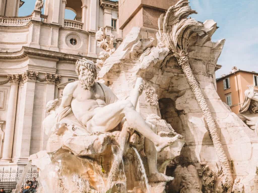 A close-up of the Fontana dei Quattro Fiumi in Piazza Navona, Rome, featuring the muscular sculpture of the river god representing the Rio de la Plata, with detailed rock formations and cascading water against the backdrop of Sant'Agnese in Agone church