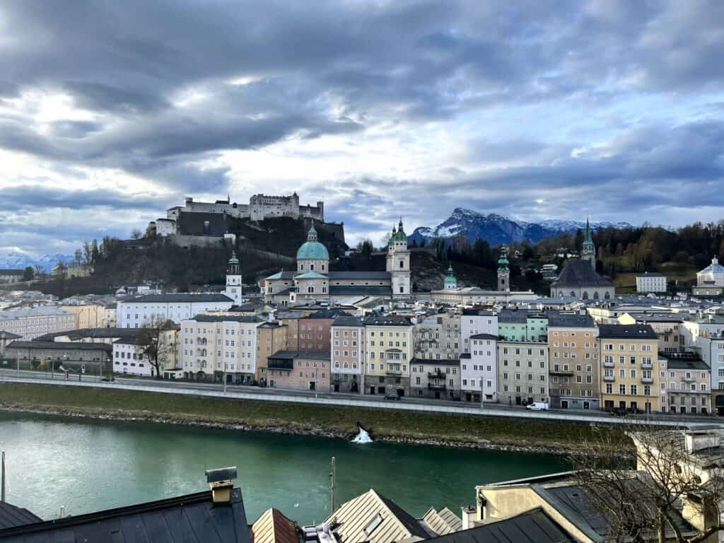 Panoramic view of Salzburg, Austria, with the Hohensalzburg Fortress atop the small hill, surrounded by historic baroque architecture under a dramatic sky. The Salzach River flows in the foreground, with the Eastern Alps rising in the background.