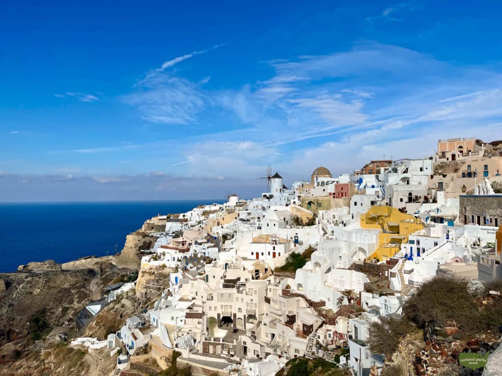 Cliffside view of Oia, Santorini, with its distinctive white-washed buildings and blue domes, nestled on rugged volcanic cliffs above the Aegean Sea, with a clear blue sky above and traditional windmills accentuating the horizon