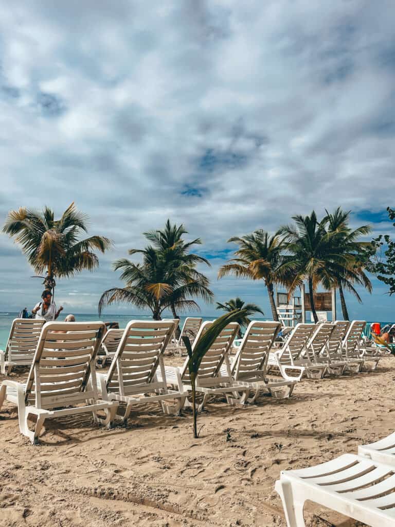 Row of empty white lounge chairs on the sandy beach of Tabyana Beach Club, Roatan, under a sky streaked with clouds, with tall palm trees swaying gently in the background and a glimpse of the serene sea beyond