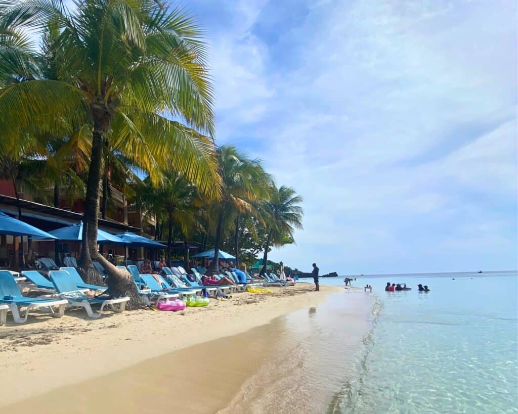West Bay Beach in Roatan lined with sun loungers under blue umbrellas, with visitors relaxing under the shade of tall coconut palms by the water's edge, as others wade into the inviting, clear Caribbean Sea.