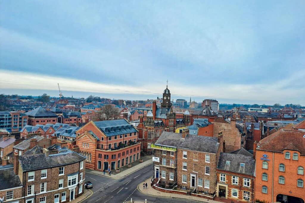 Elevated view of York, England, showcasing its mix of historic and modern architecture, with the prominent clock tower of the York Art Gallery, terracotta rooftops, and a construction crane in the distance against a soft blue sky.