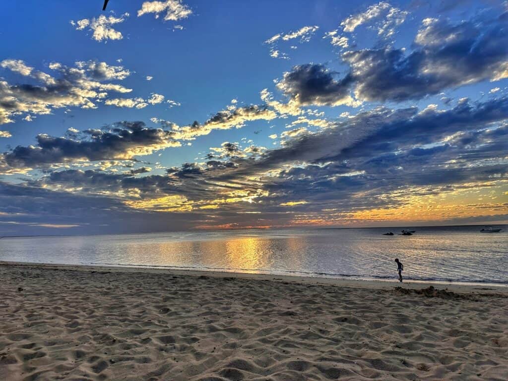 A serene sunset on a Fijian beach, with the sky painted in hues of blue, gold, and orange as clouds are illuminated by the waning sun, reflecting on the calm sea where a few boats are anchored, while a lone person walks along the shore