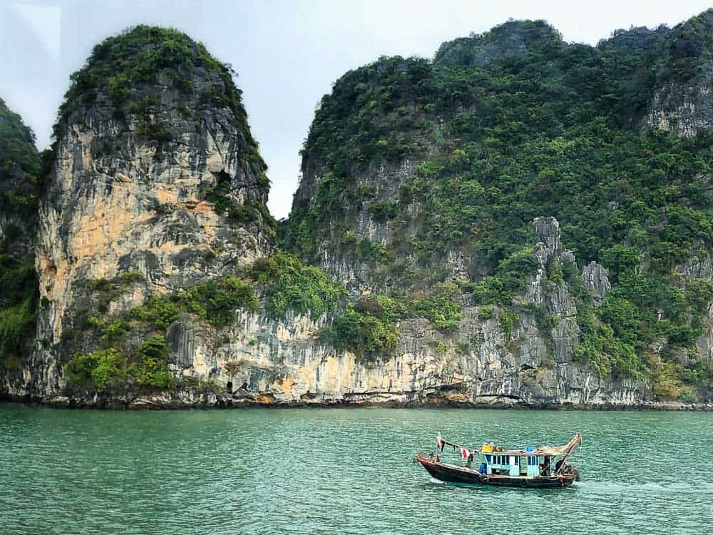 A traditional Vietnamese boat sails through the emerald waters of Ha Long Bay, with towering limestone karsts covered in lush greenery in the background