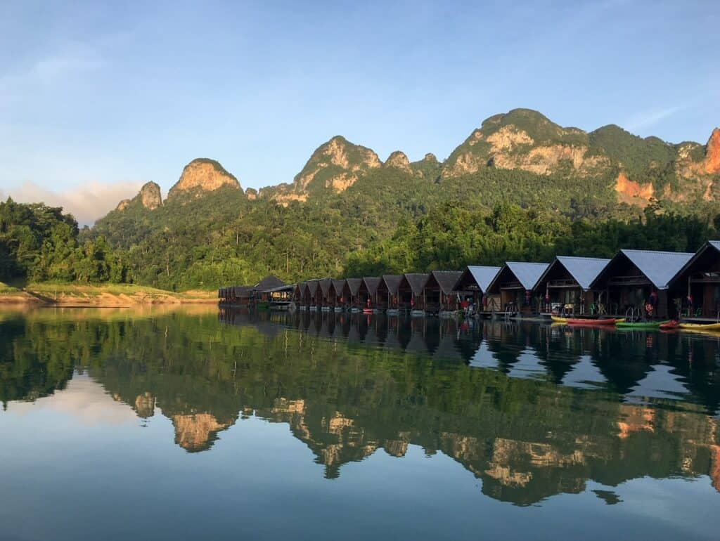 Floating bungalows on the tranquil waters of Cheow Lan Lake in Khao Sok National Park, Thailand, with a backdrop of lush, rugged limestone mountains reflecting on the glassy surface during a serene morning.