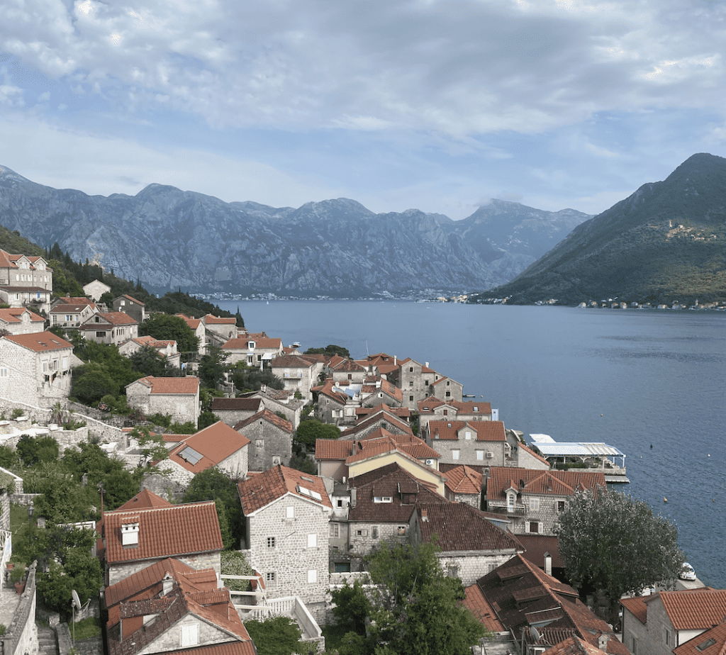 The image features a stunning view of Kotor, Montenegro, showcasing the historic old town with its traditional stone houses and terracotta roofs nestled against the calm waters of the Bay of Kotor. The dramatic backdrop of the rugged Dinaric Alps completes this picturesque scene, typical of the Adriatic coastline.