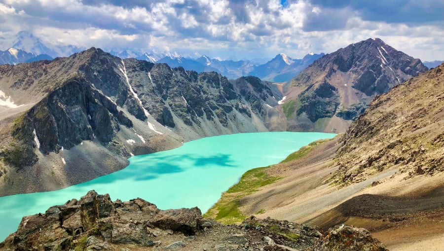 The breathtaking Ala-Kul Lake in Kyrgyzstan, nestled among rugged mountain peaks with its striking turquoise waters creating a stark contrast against the gray rocky landscape under a partly cloudy sky