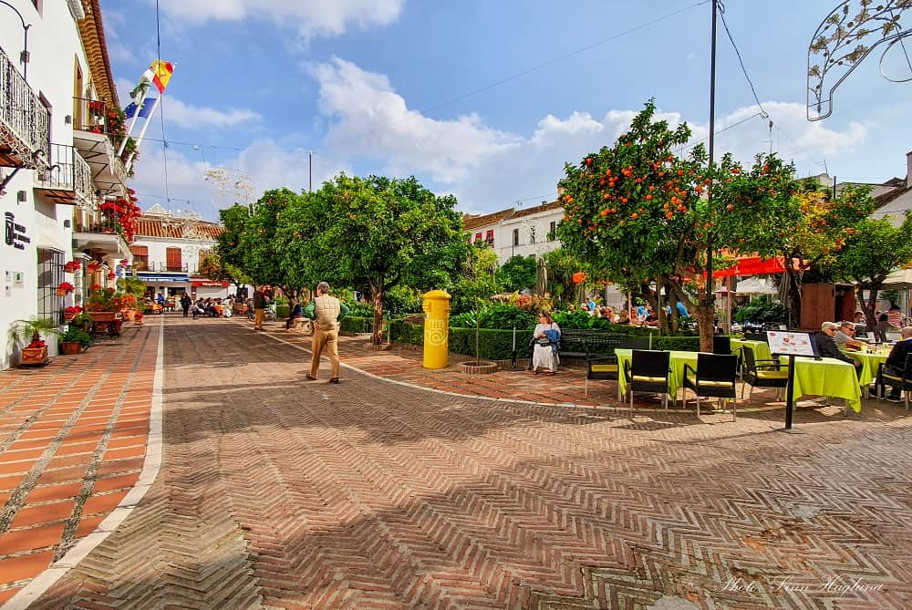 This image shows a vibrant street scene in Marbella, Spain. The foreground features a patterned brick road with people walking and a man in a beige jacket. The midground is lively with outdoor dining under orange trees in full bloom. White buildings with balconies adorned with red flowers, and a clear blue sky with a few clouds complete the background.