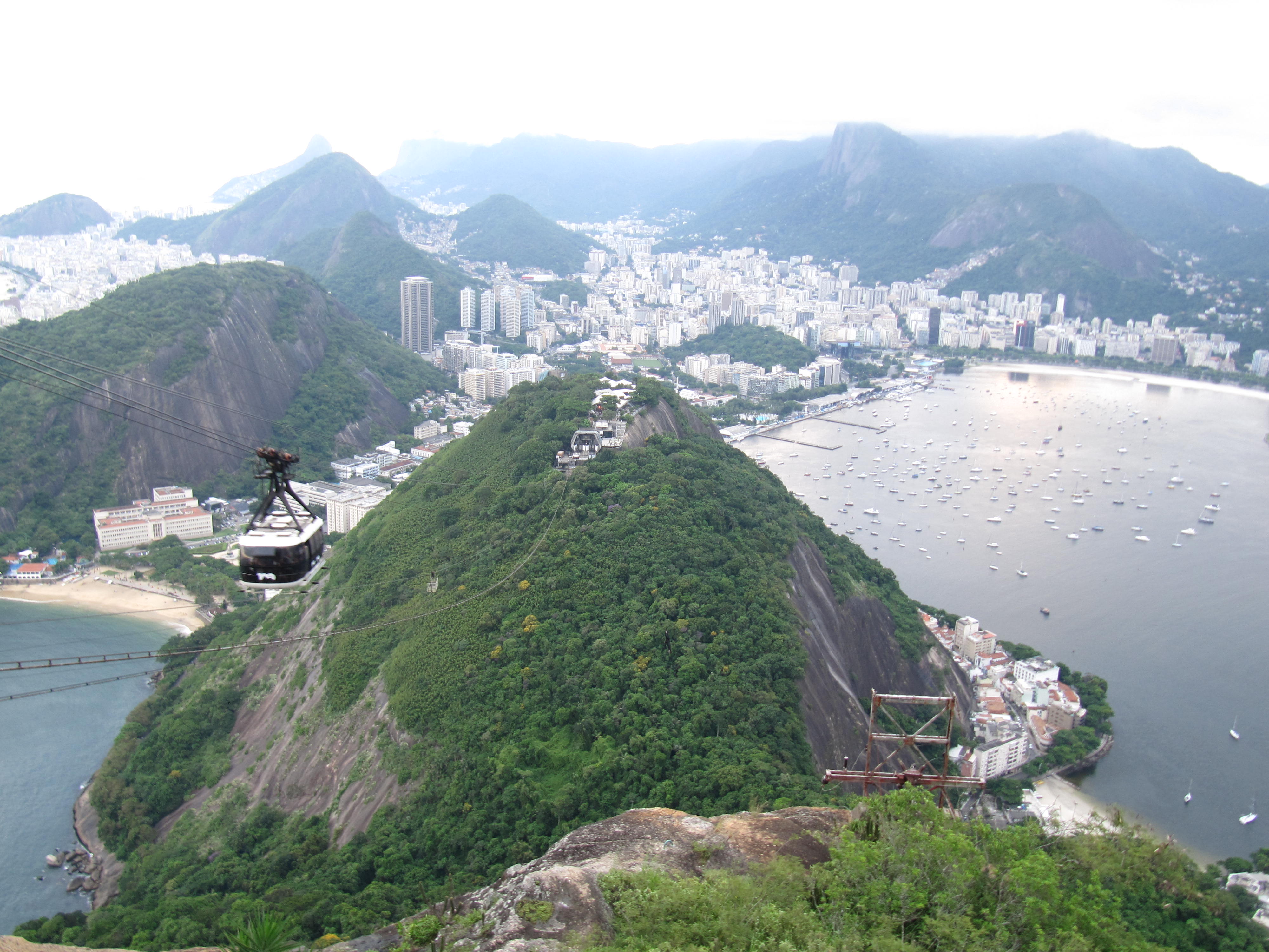 Aerial view of Rio de Janeiro from Sugarloaf Mountain, with the iconic cable car ascending the peak, overlooking the densely populated cityscape, the sprawling Botafogo Bay filled with boats, and the distant Corcovado Mountain