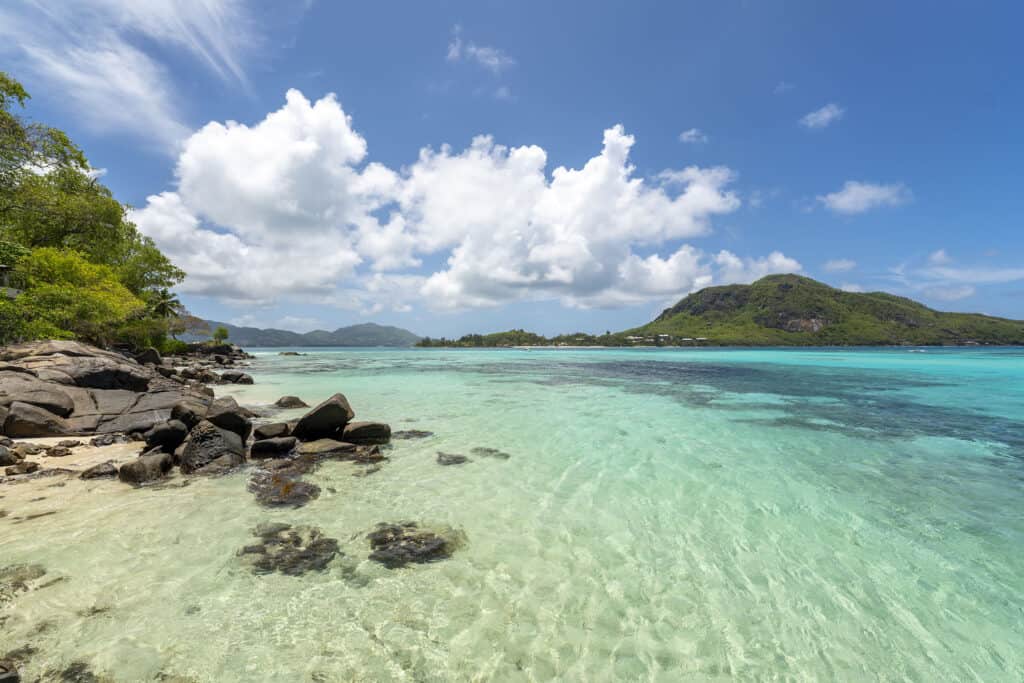 Crystal-clear turquoise waters of the Seychelles with a rocky shoreline covered in lush greenery, overlooking Round Island, under a blue sky with majestic cumulus clouds.
