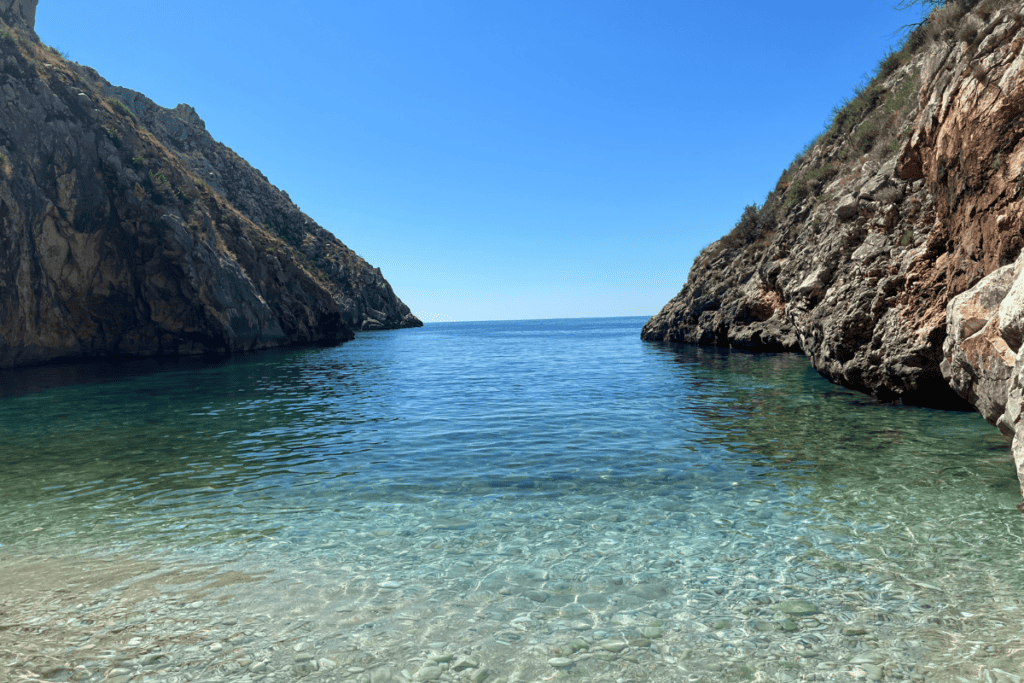 A hidden cove with crystal-clear waters in Albania, flanked by rugged cliffs that gently slope into the Adriatic Sea, under a clear blue sky on a bright sunny day.