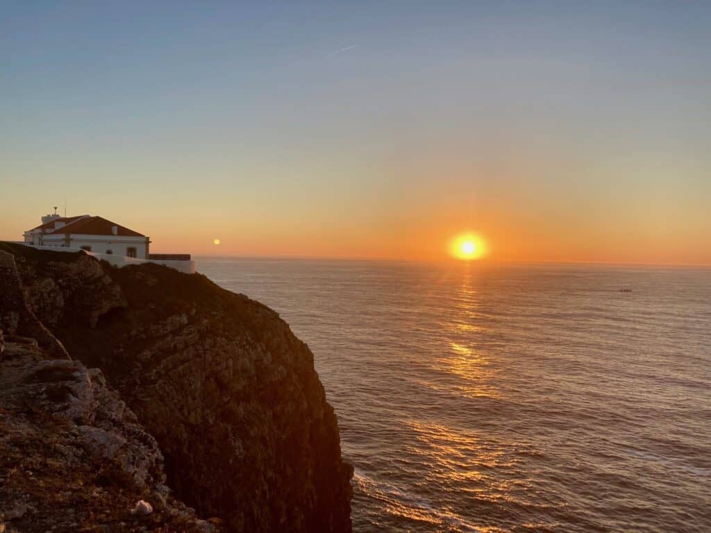 The image captures a breathtaking sunset view at the Algarve coast in Portugal, with the sun dipping into the ocean horizon. A white building perches on the edge of a rugged cliff, overlooking the vast sea that reflects the warm hues of the sunset, enhancing the tranquil and picturesque scenery.