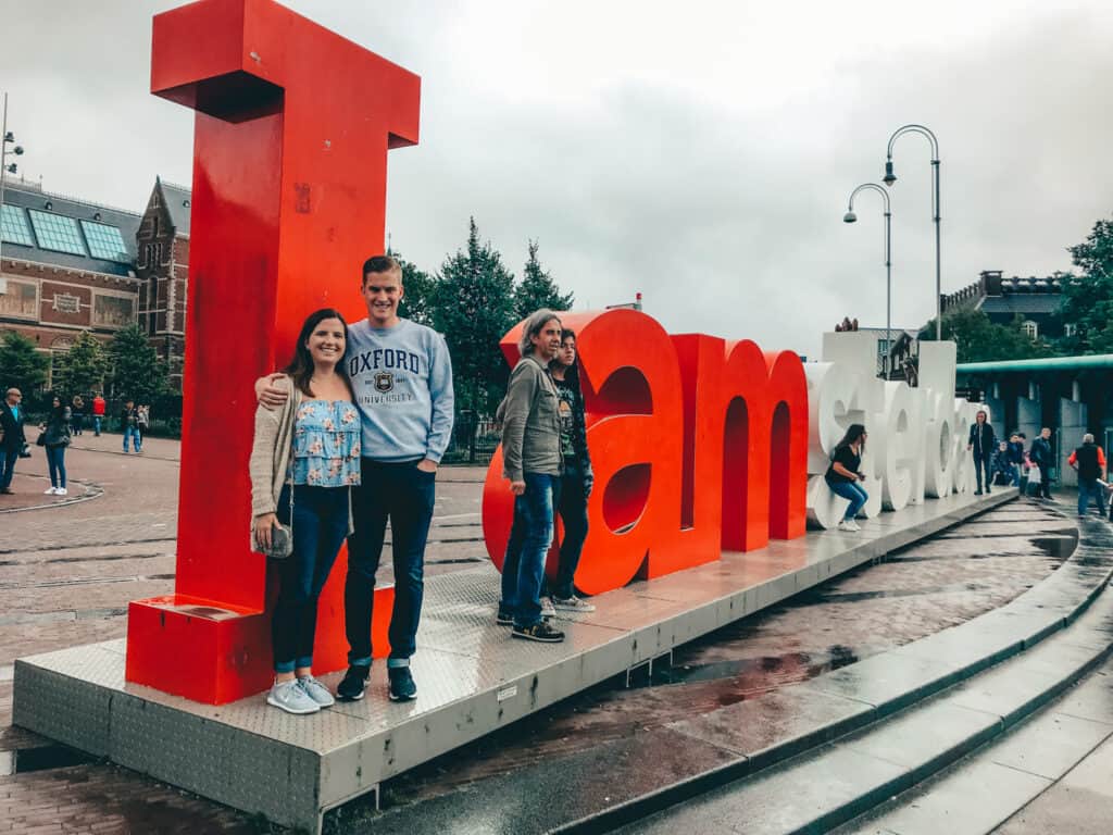 The image shows three people posing in front of the iconic 'I amsterdam' sign, with two standing by the 'I' and one by the 'd'. The large red and white letters of the sign create a striking contrast against the cloudy sky, and the busy surroundings suggest a popular tourist spot. The wet ground indicates recent rain, adding to the lively ambiance of the scene.