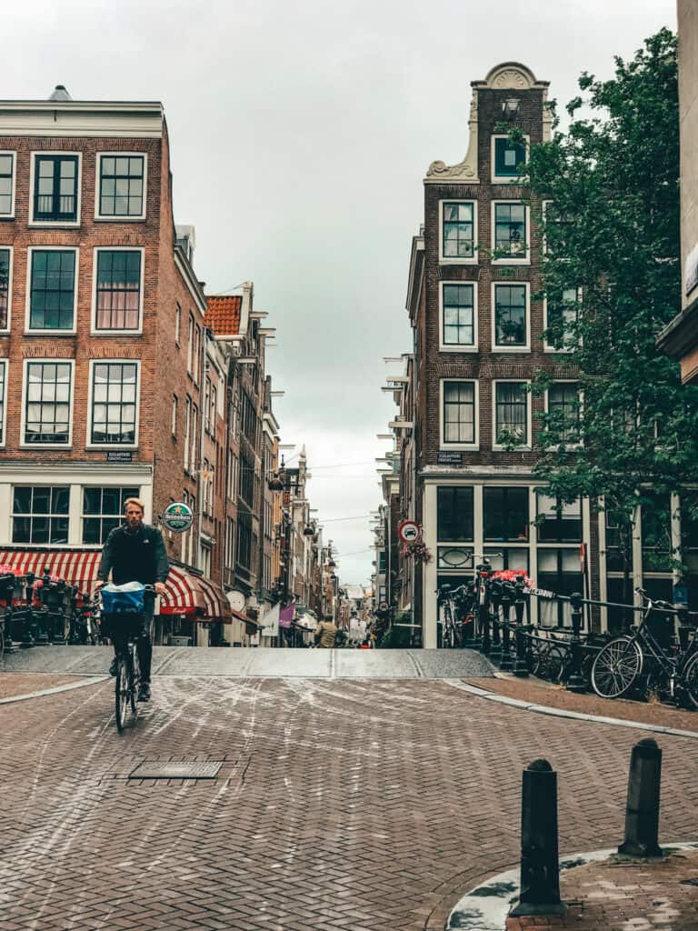 The image depicts a typical Amsterdam street lined with historic Dutch buildings featuring gabled facades. A cyclist is captured in the foreground, riding on the cobblestone street that reflects a common mode of transportation in the city. The overcast sky suggests a cloudy day in this urban, European setting.