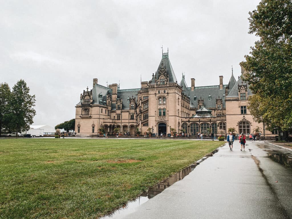 Visitors strolling near the historic Biltmore Estate in Asheville, North Carolina on a cloudy day. The grand mansion's French Renaissance architecture, complete with turrets and ornate detailing, stands out against the overcast sky