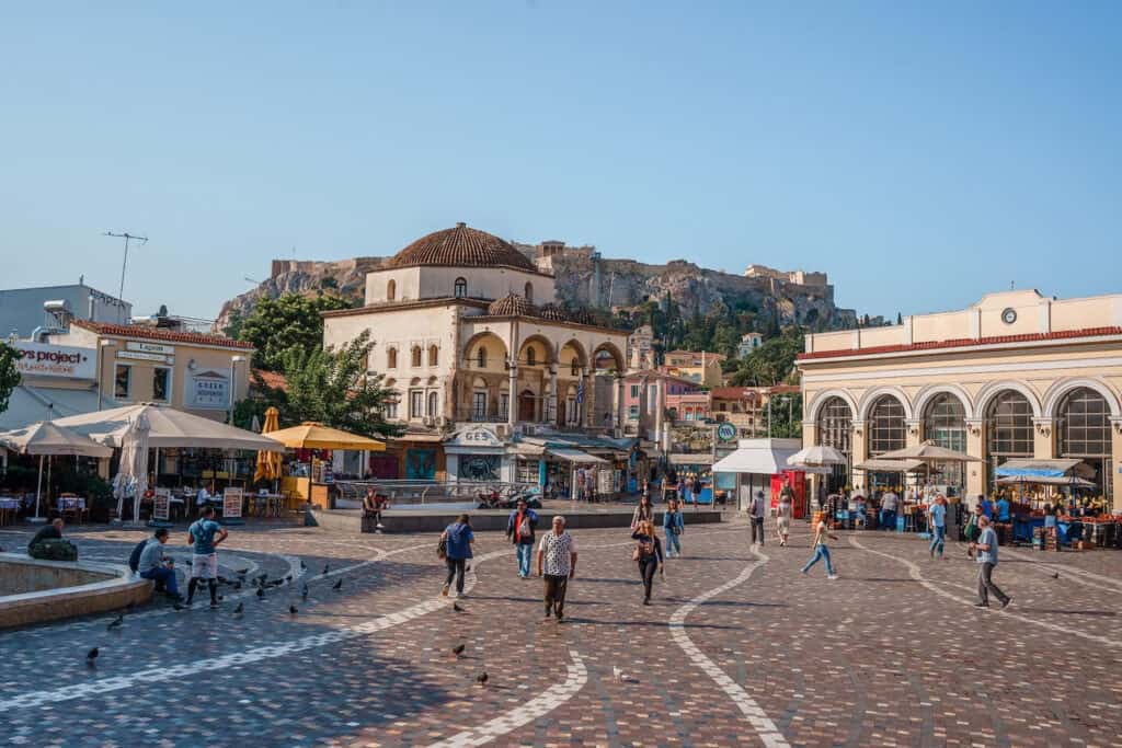 The image displays a lively town square in Athens, Greece, with pedestrians walking across the patterned pavement. The square is flanked by neoclassical buildings, cafes with outdoor seating, and a Byzantine-style church, with the Acropolis visible in the distant background under a clear blue sky.
