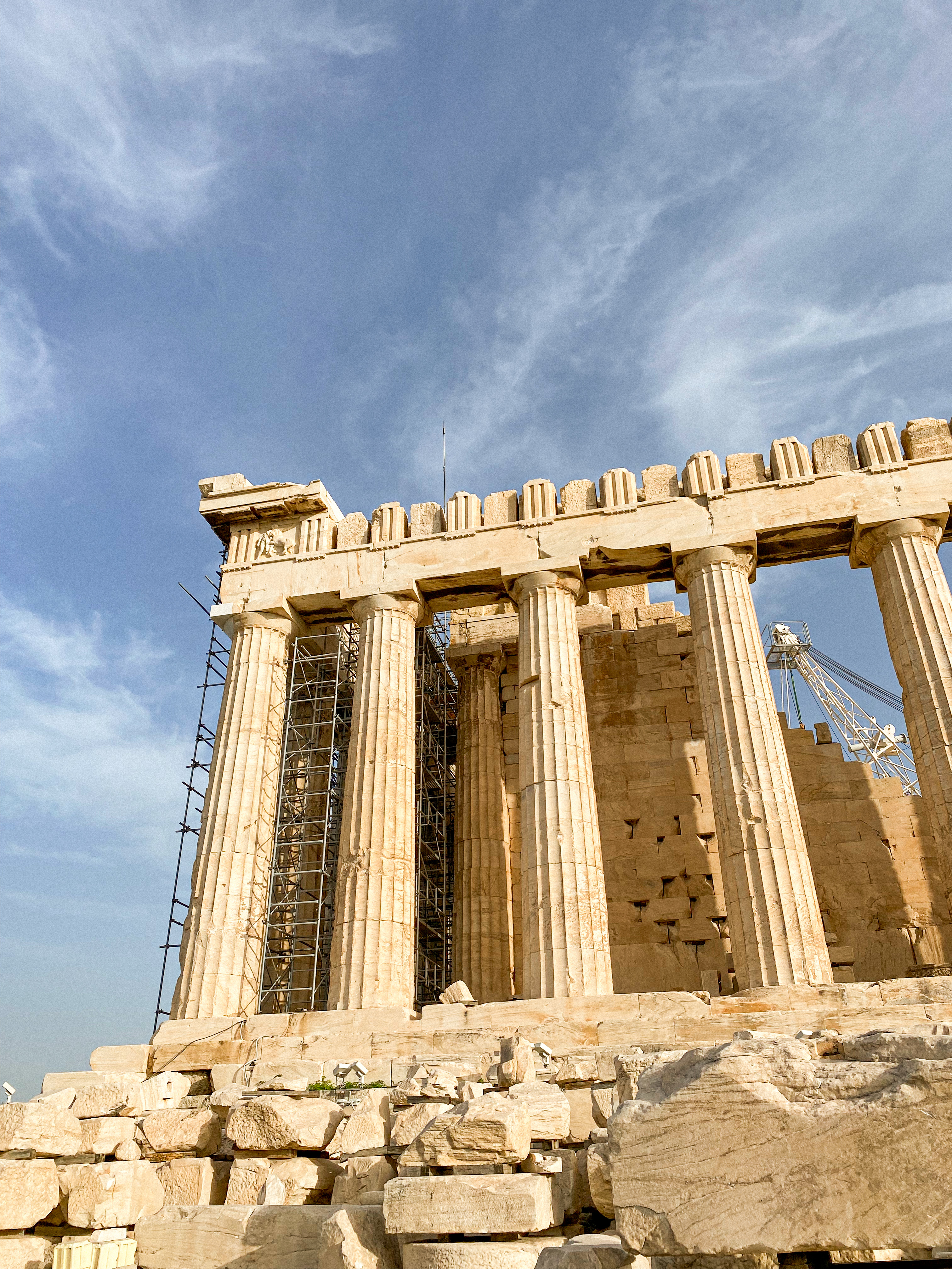 The Parthenon under a clear blue sky, showcasing the ancient structure's iconic Doric columns and ongoing restoration work, with scaffolding visible against the sun-bleached stone on the Acropolis of Athens