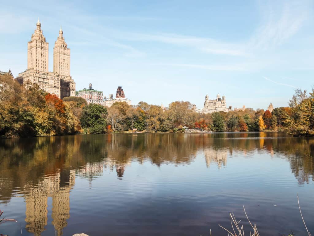 Autumn foliage frames the serene waters of The Lake in Central Park, New York City, with the iconic towers of the San Remo apartments reflecting in the calm water.