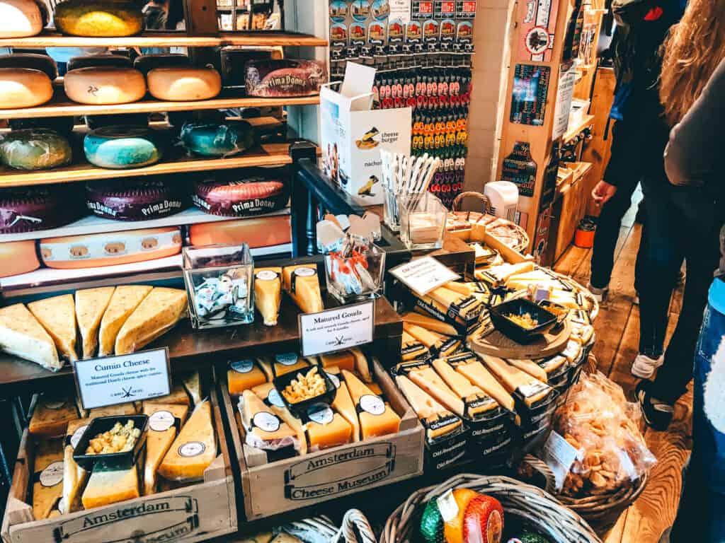 The image captures the interior of a vibrant cheese shop, possibly part of a cheese museum, filled with an array of colorful cheese wheels on shelves and cut cheese samples available for tasting. Signs like 