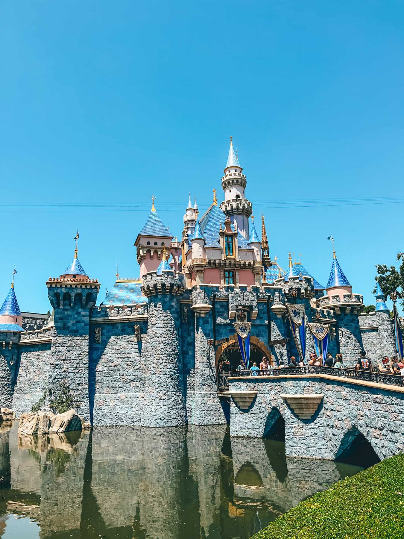 Cinderella's Castle at Disneyland, with its iconic pink and blue turrets, reflected in the moat under a clear blue sky. The fantasy architecture and vibrant banners create a magical atmosphere in the theme park.