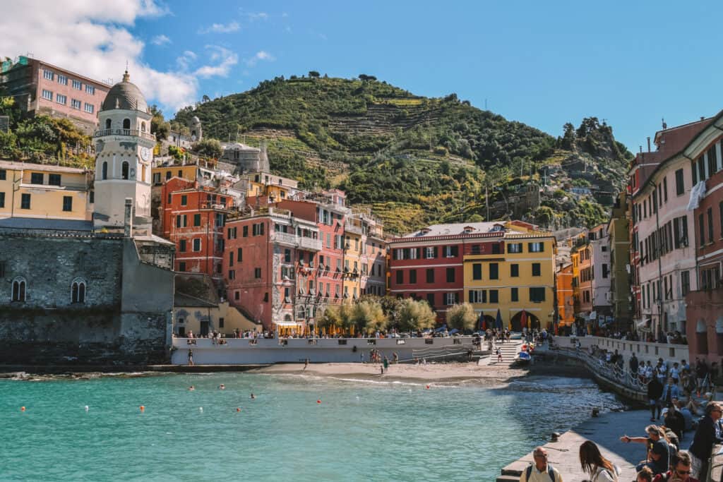 The image captures the vibrant and picturesque setting of Cinque Terre, Italy. Colorful houses and buildings cling to the steep terraced hillsides above the small beach and turquoise waters, with a bustling waterfront promenade beneath a clear blue sky.