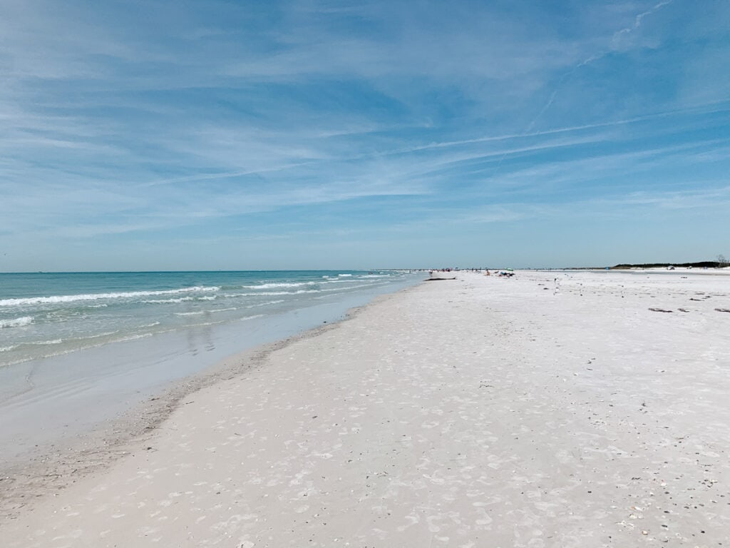 Expansive view of Clearwater Beach in Florida with its soft white sands and gentle surf. The beach extends towards the horizon under a vast blue sky with wispy clouds, highlighting the serene and sunny coastal atmosphere.