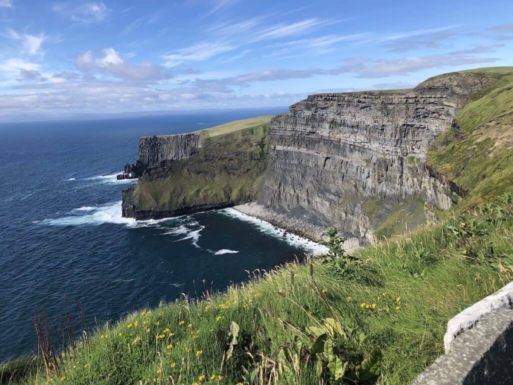 The Cliffs of Moher rise majestically above the Atlantic Ocean, with layers of limestone visible along their heights, the sea's waves gently crashing against the base, all under a bright sky on the west coast of Ireland.