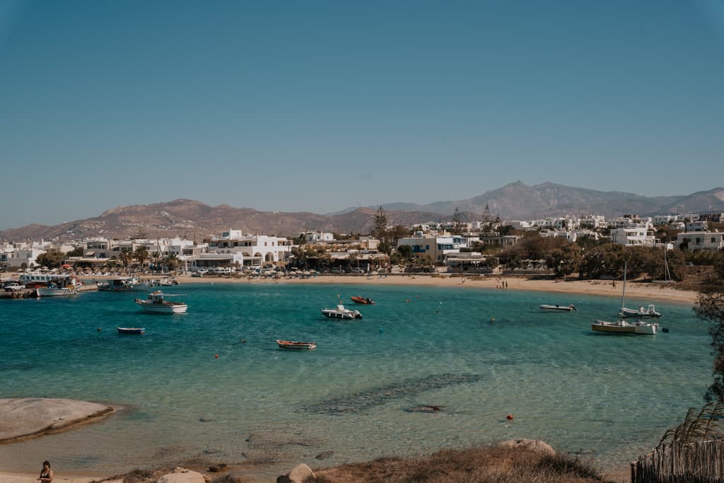 The image depicts a tranquil beach scene on one of the Cycladic Islands in Greece, with crystal clear blue waters and a variety of boats moored near the shore. The background shows a stretch of typical Cycladic architecture with white buildings against a backdrop of rugged hills under a clear blue sky.