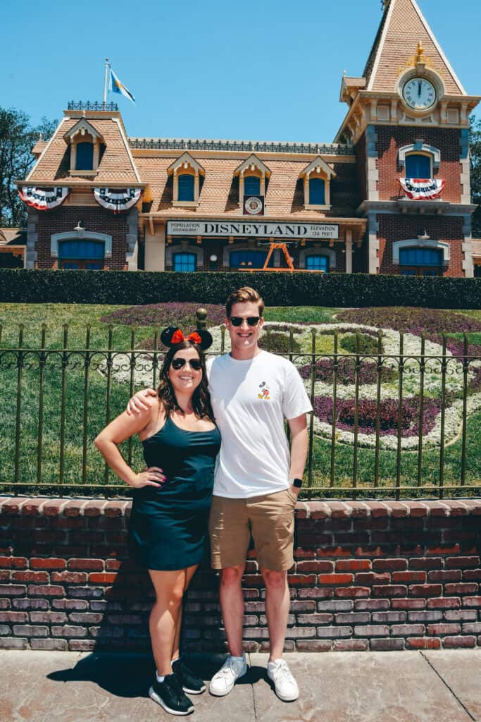 A smiling couple stands in front of the Disneyland entrance under a clear blue sky. The woman is wearing mouse ears and the man is in a white t-shirt with a Disney character. In the background, the iconic Disneyland station building is adorned with American flags and a floral display spelling 'Disneyland